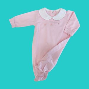 Pink baby onesie with a white Peter Pan collar laid on a turquoise background, long sleeves and delicate embroidery along the chest area