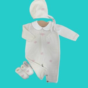 White knitted baby outfit set (hat, dress, booties) with small pink rose embroidery on a teal background.