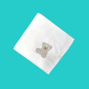 White quilted baby blanket folded at the corner with a small brown teddy bear embroidery, on a teal background